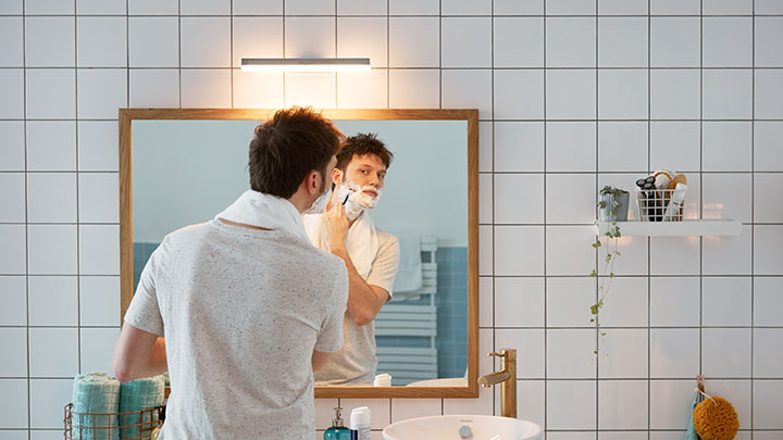 Mężczyzna golący się przed lustrem łazienkowym oświetlonym przez zamontowane na ścianie oświetlenie Philips LED